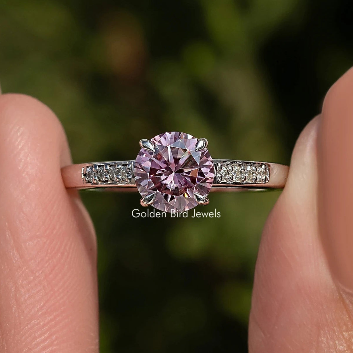 Pink gemstone ring held between two fingers with a blurred green background