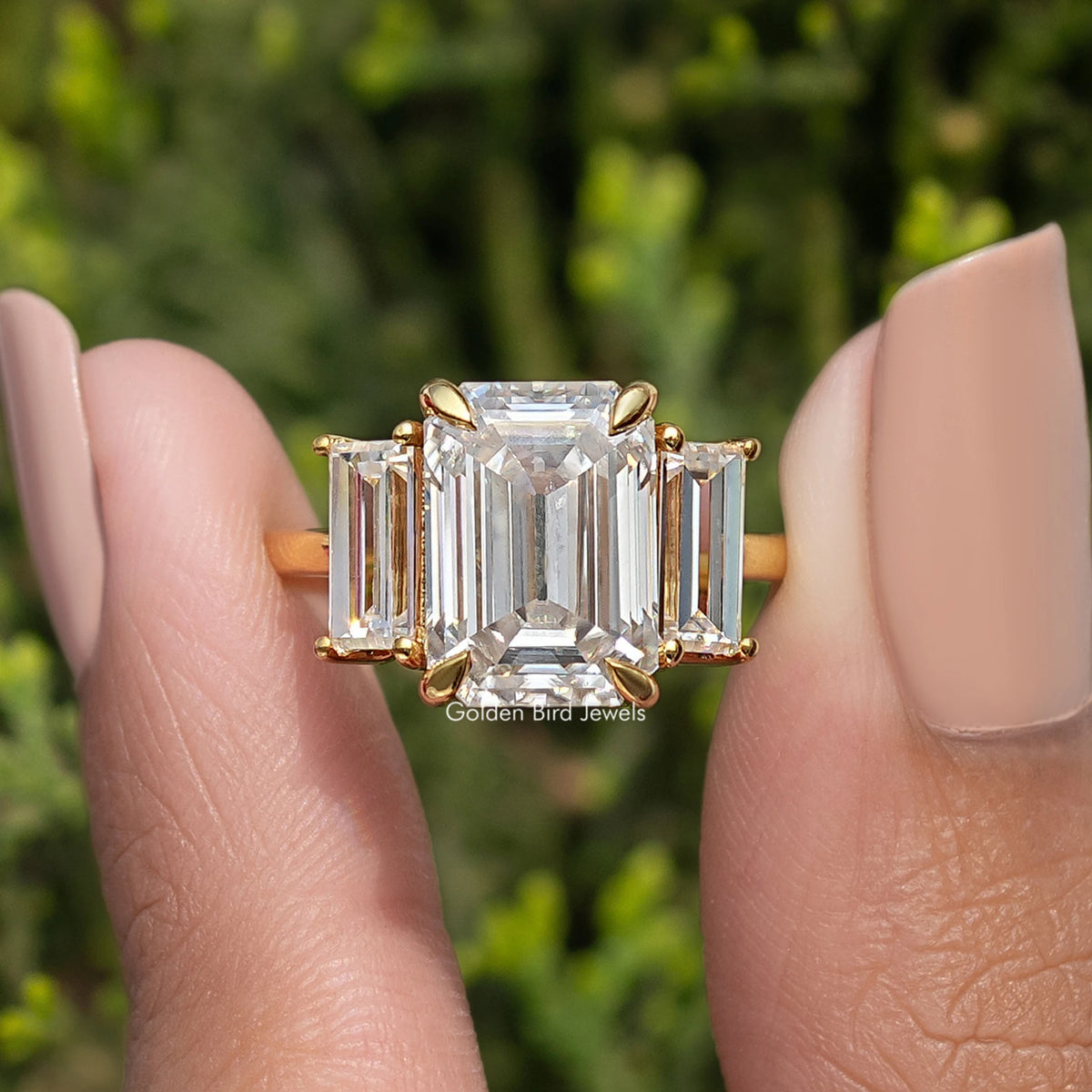 Three-stone emerald-cut moissanite ring with square center stone and side stones, gold-tone band.